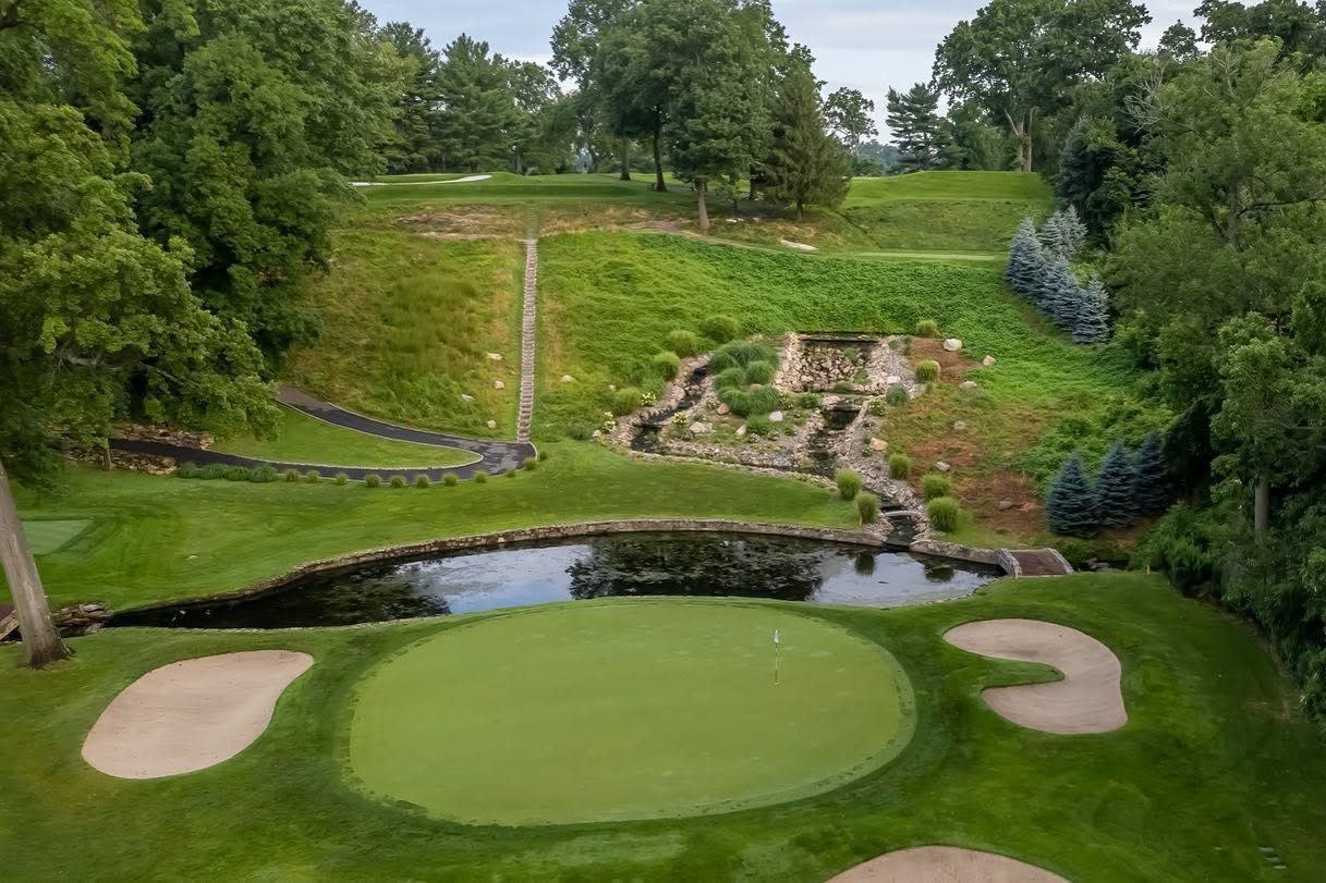 Golf course green with water feature and sand traps. Green grass, trees, and cloudy sky.