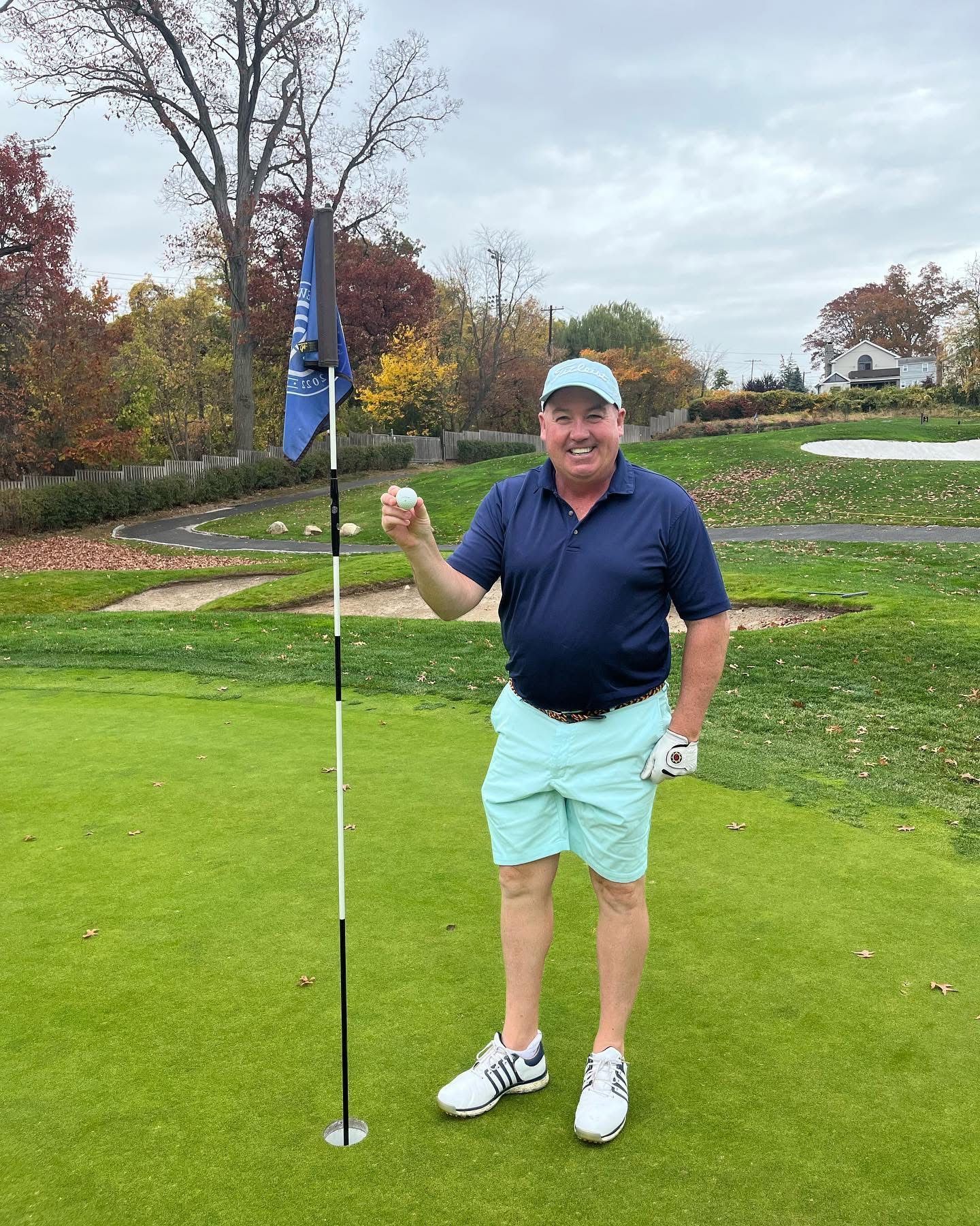 Man holding golf ball near the hole on a golf course; wearing blue and light green clothing.