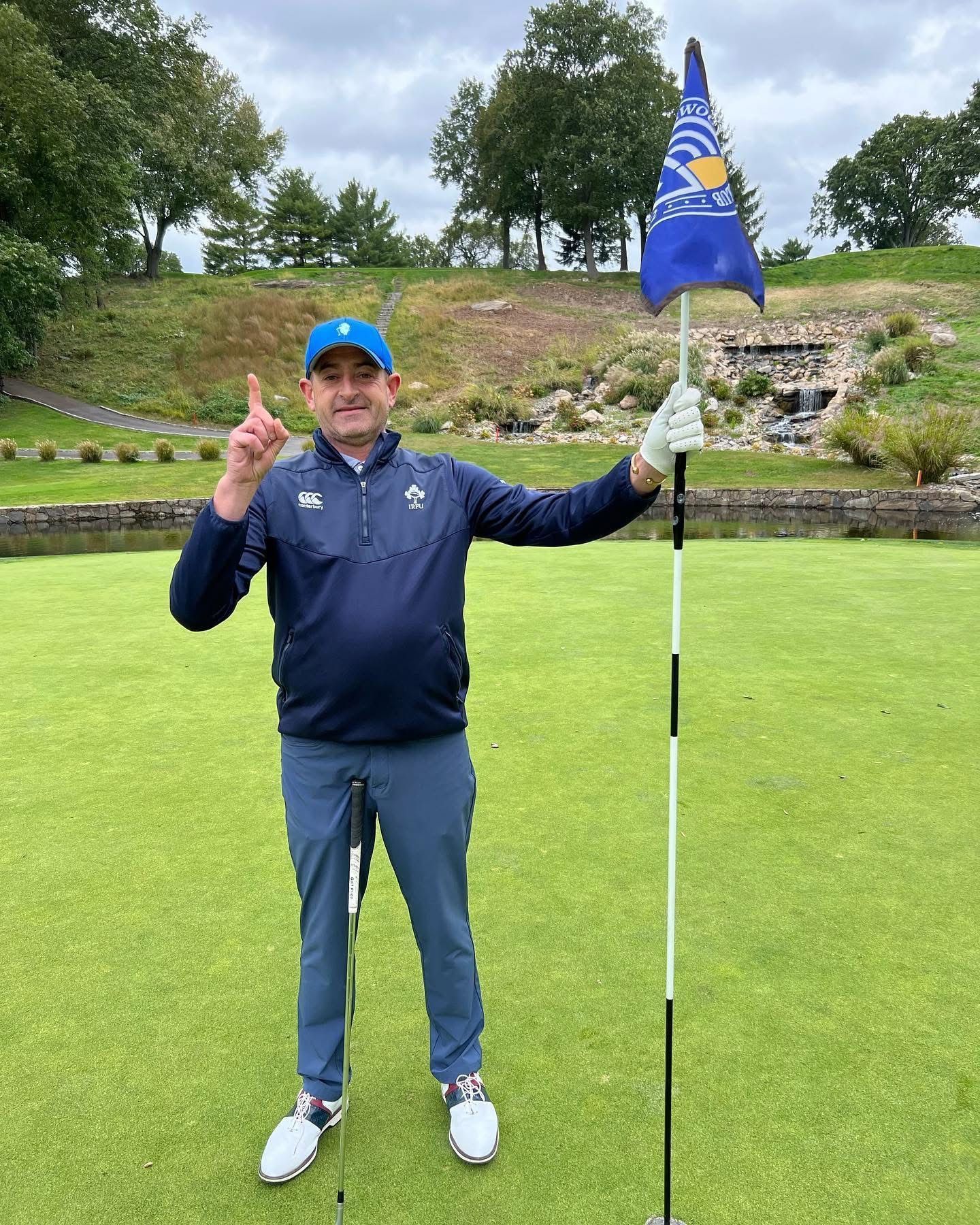 Golfer celebrating on the green, holding flag. Wearing blue, pointing up, with background of waterfall and trees.