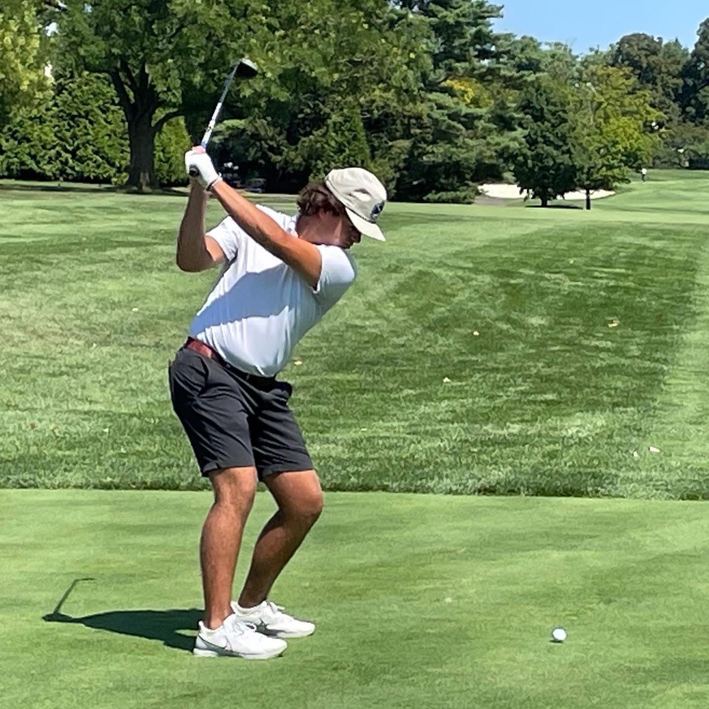 Golfer swinging a club on a green course. Wearing white shirt, dark shorts, hat. Sunny day.