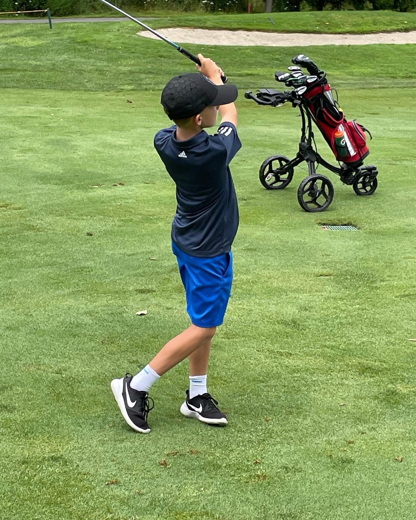 Boy in blue shorts and black shirt swings golf club on a green course.