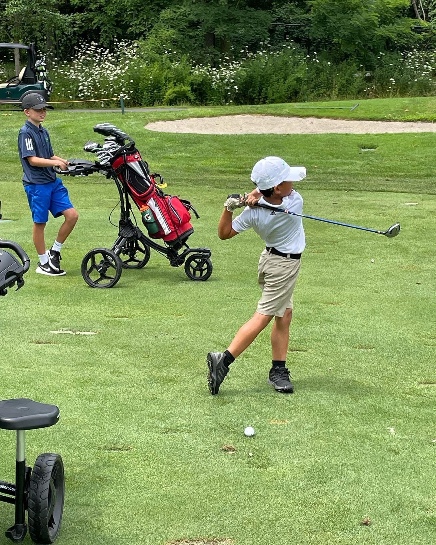 Boy in a white hat swings a golf club. Another boy stands nearby, holding a golf bag. Green grass setting.