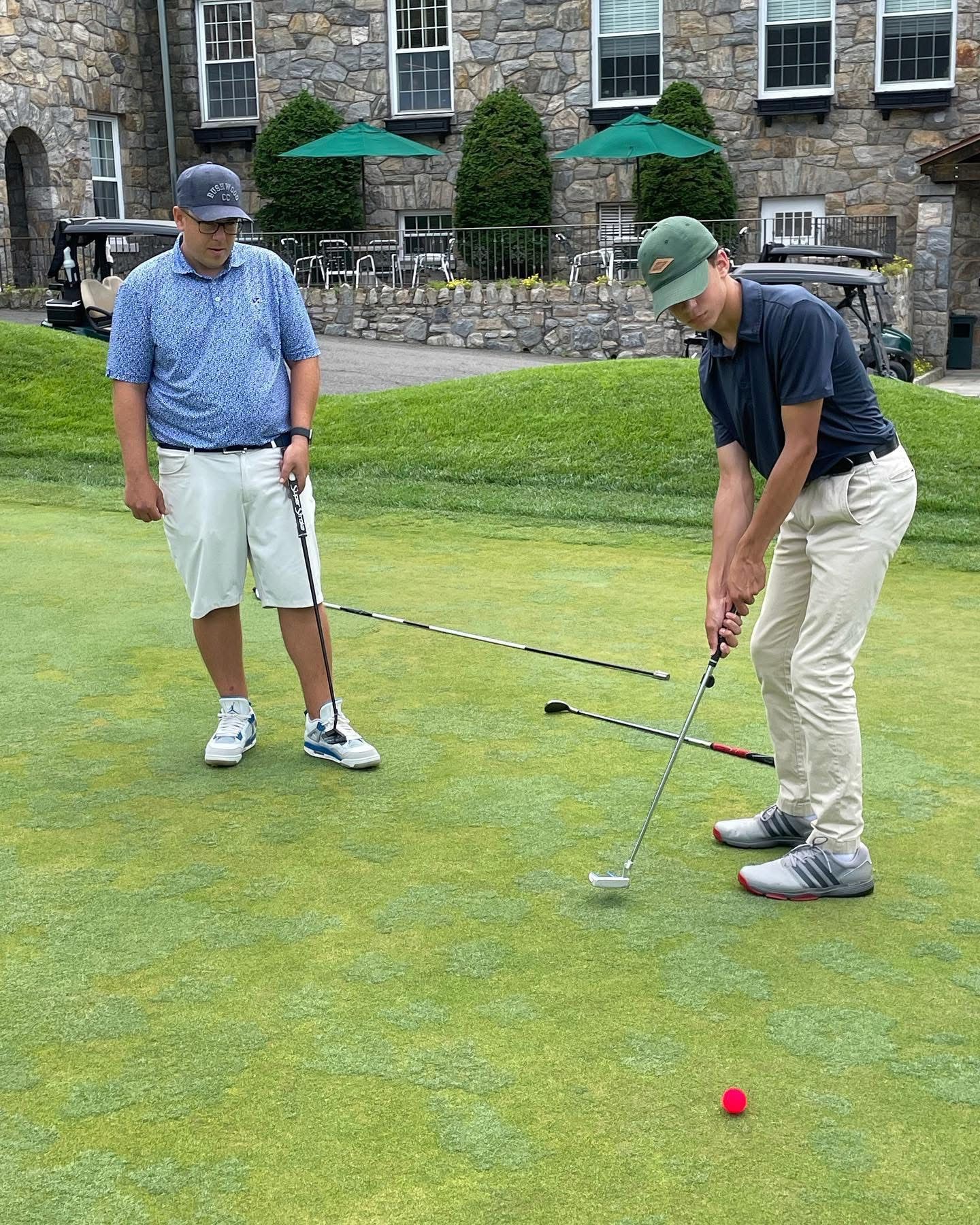 Two people putting on a golf course: one preparing to putt, the other watching. Green grass, sunny day.