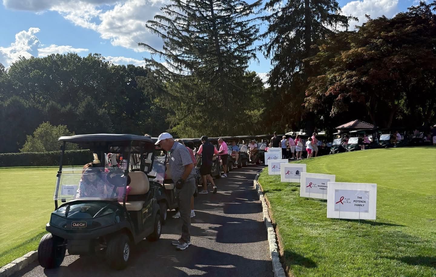 Golf carts lined up on a grassy path, people in pink clothing, signs on the lawn.