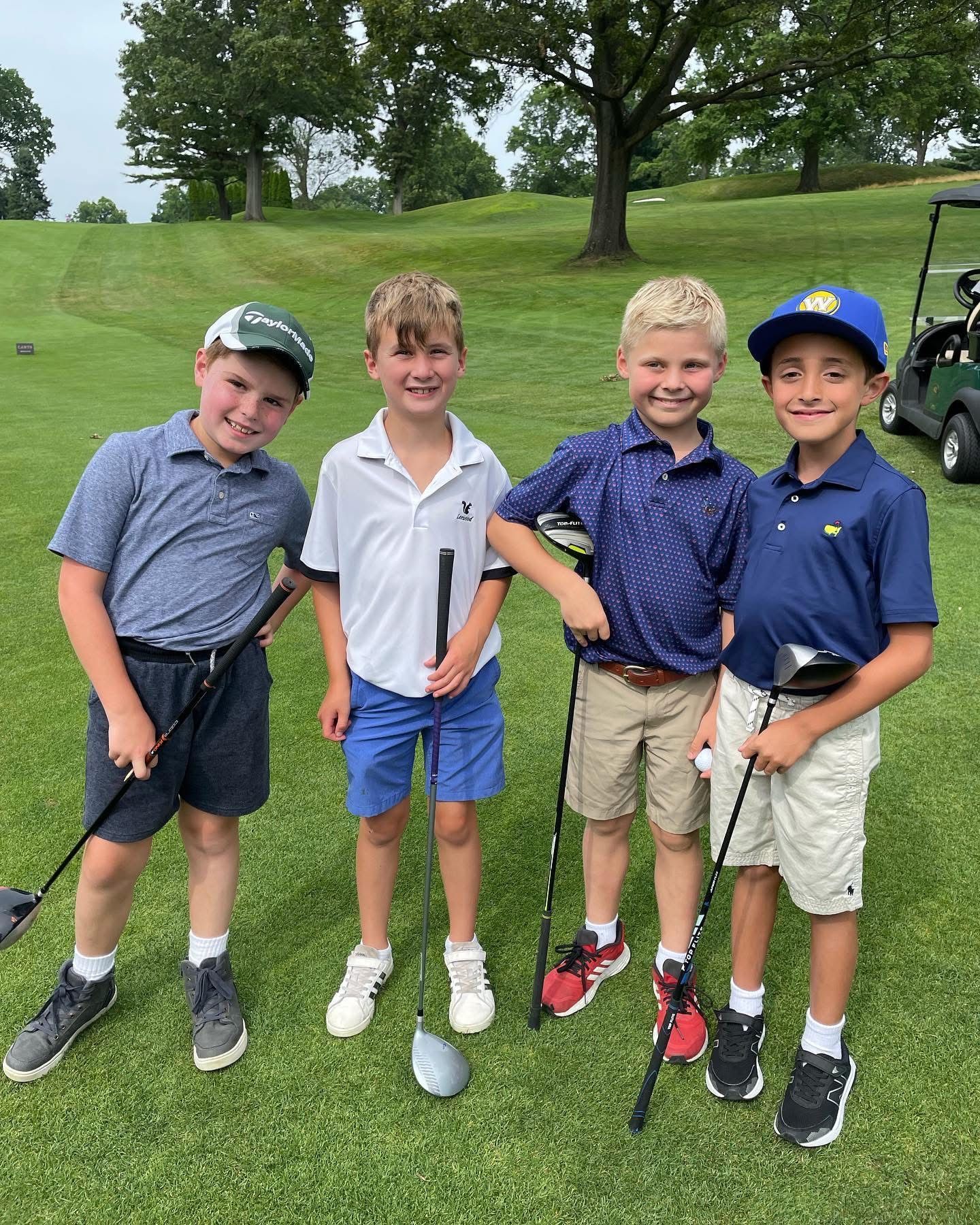 Four young boys smiling on a golf course, holding clubs. Green grass, golf cart in the background.