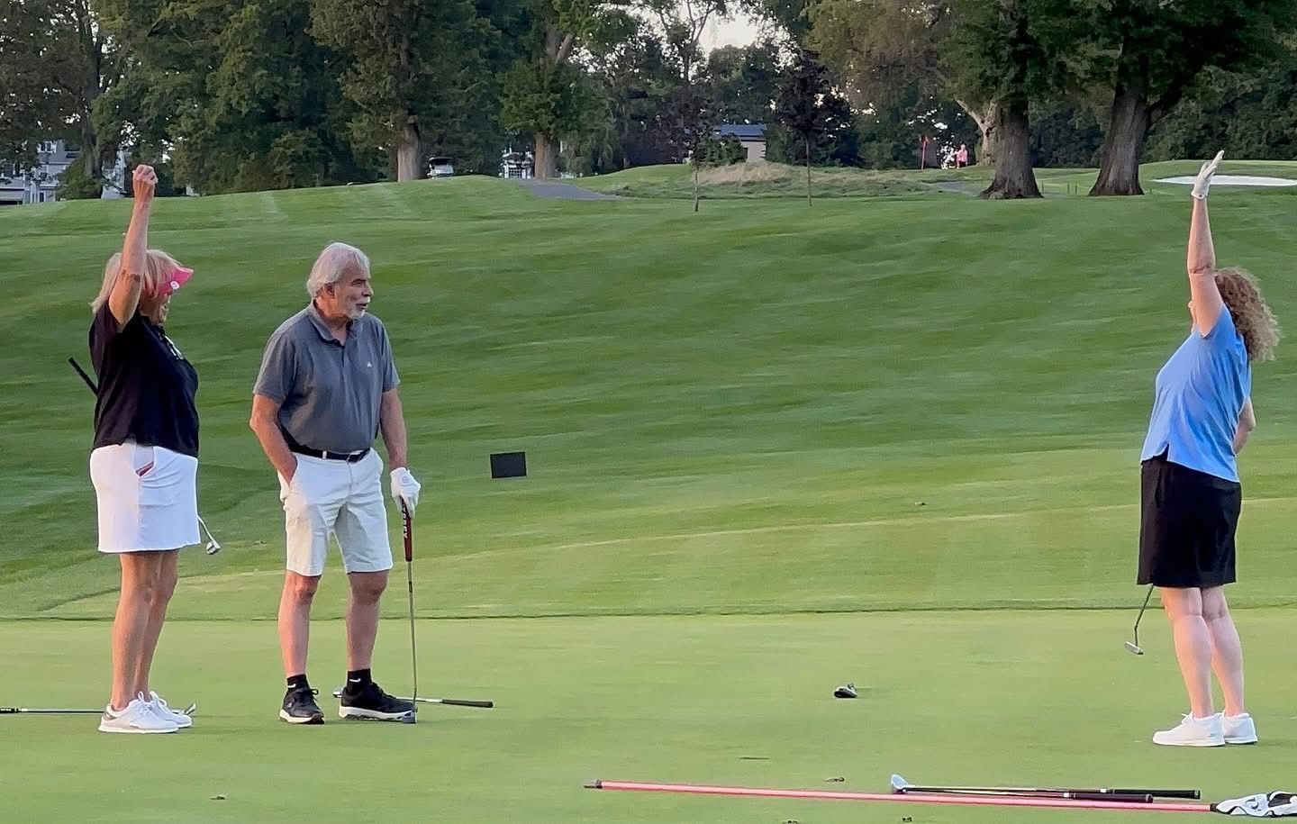 Three people on a golf course: two women with arms raised and a man with a golf club. Green grass.