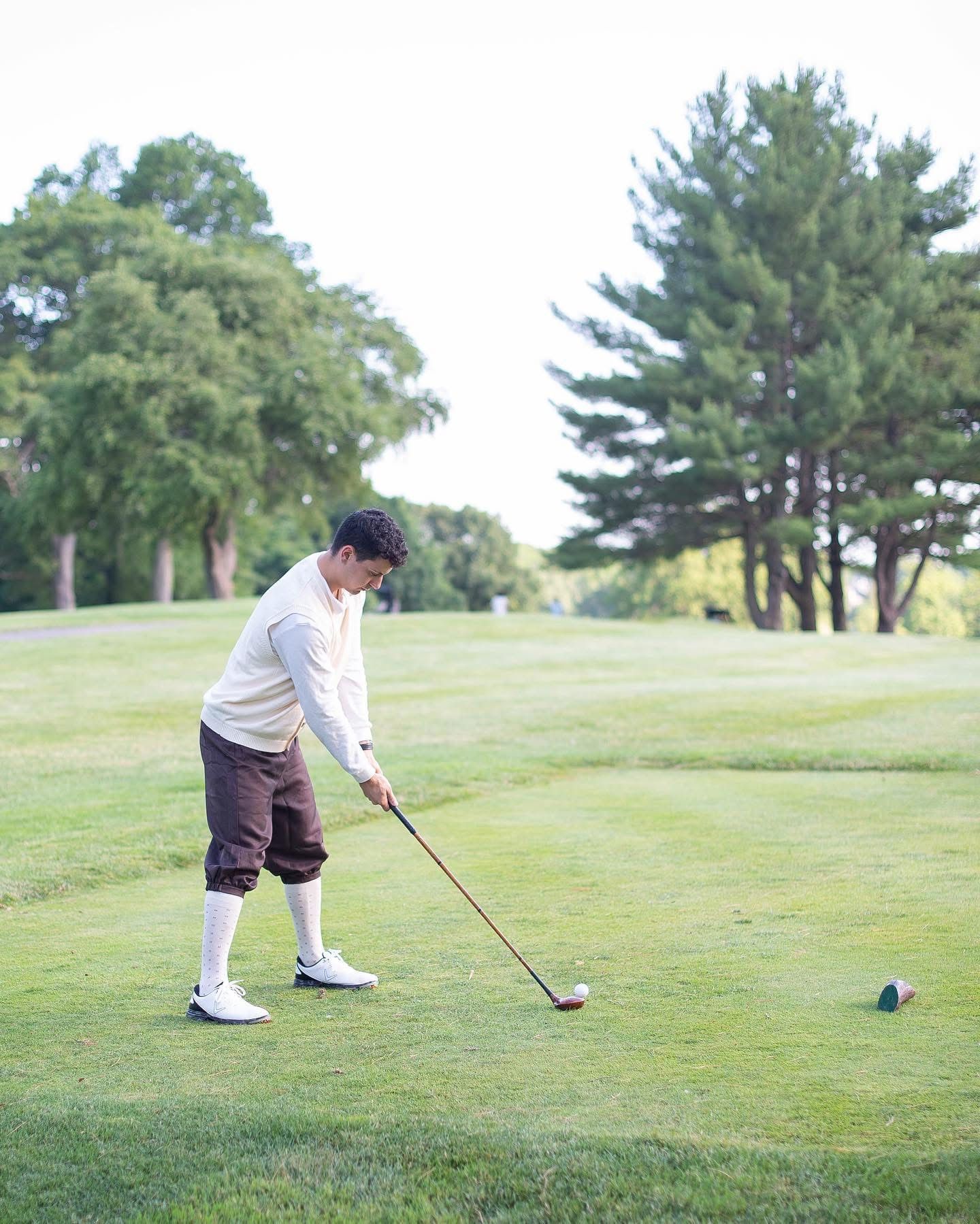 Person in vintage golf attire swings a golf club on a green course.