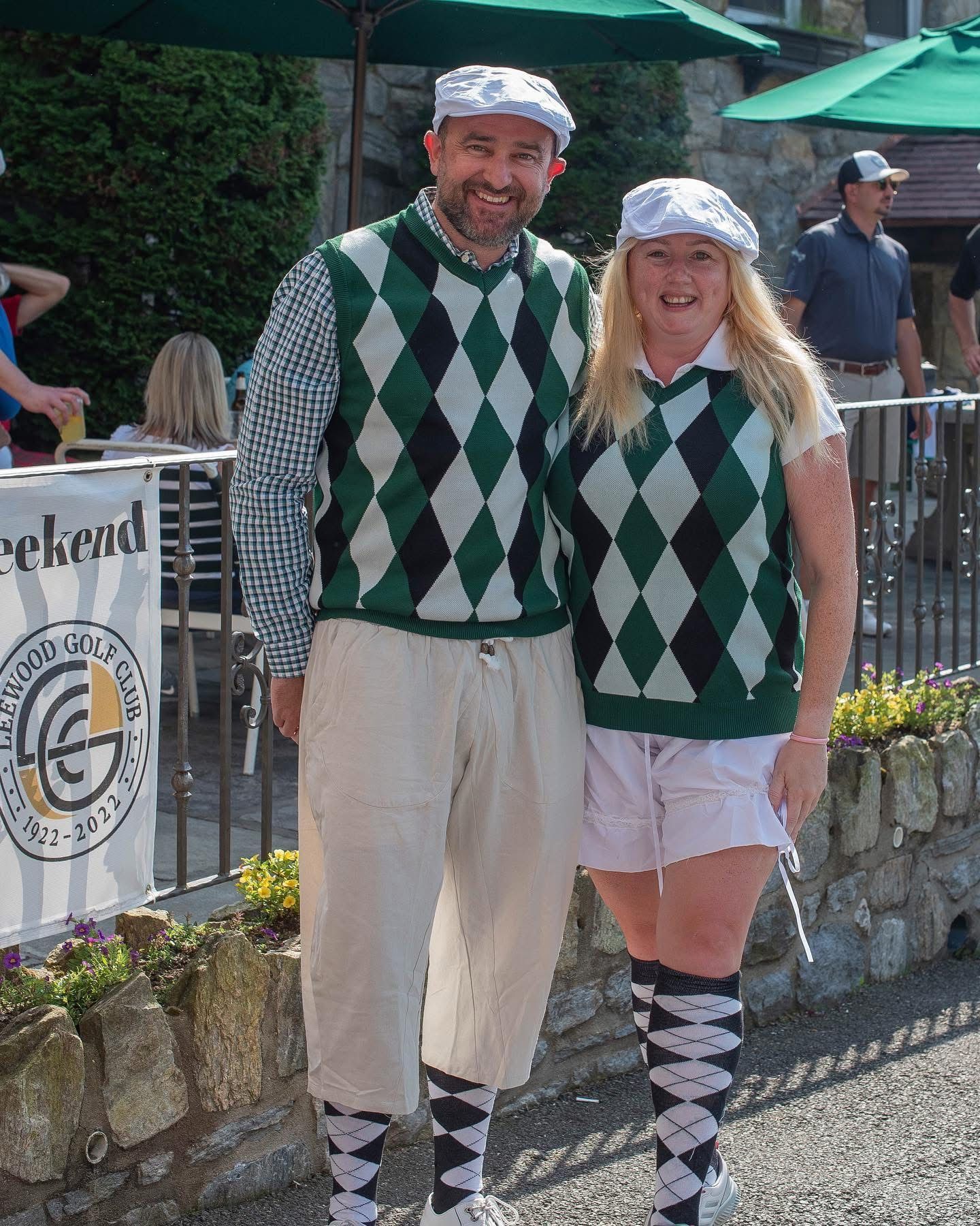 Two people in argyle vests and caps pose outdoors. Man in pants; woman in shorts, argyle socks.