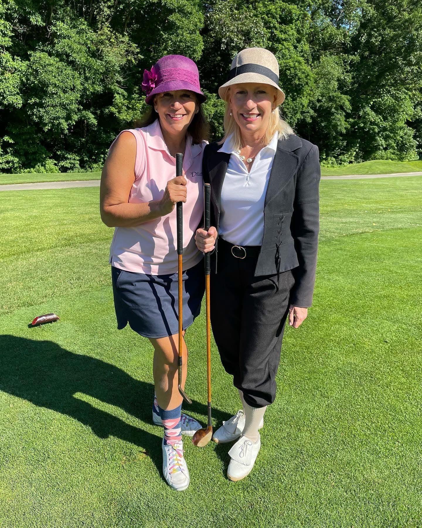 Two women in vintage golf attire on a golf course, posing with golf clubs.