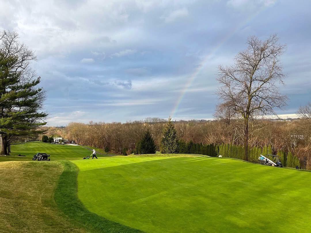 Green golf course with a partial rainbow; overcast sky. People playing golf.