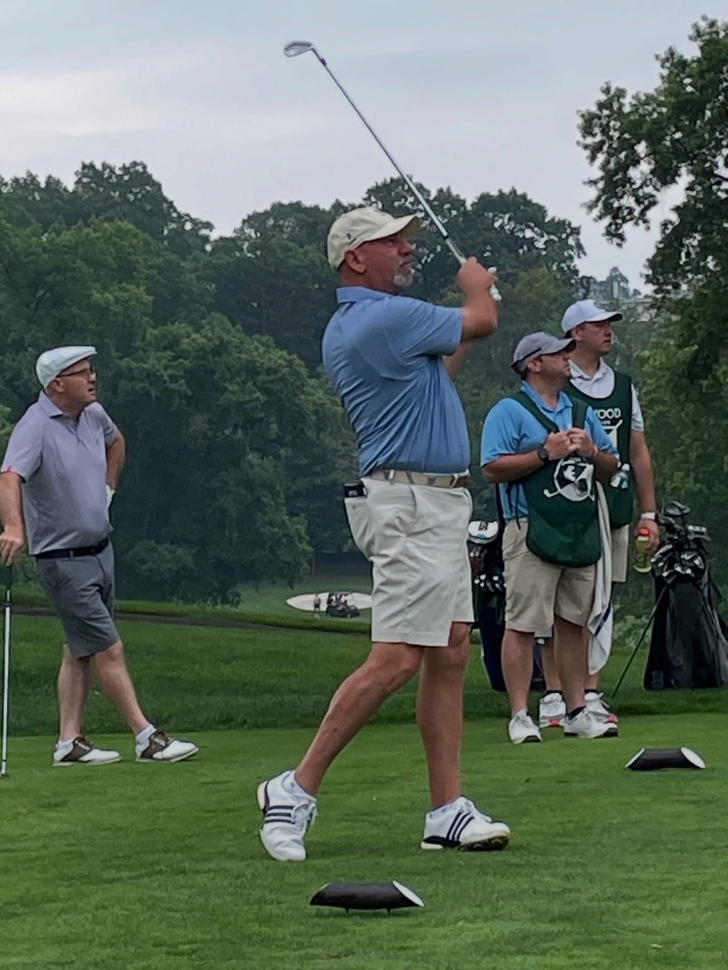 Man in light blue shirt swings golf club on a green course; spectators watch.