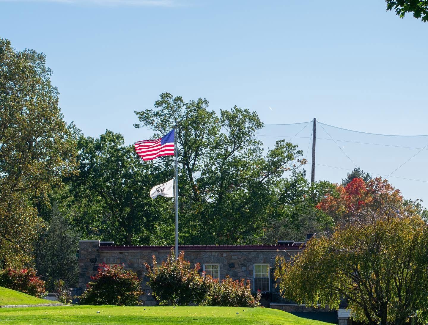 American flag and white flag on a pole near a brick wall, trees, and a golf net, on a sunny day.