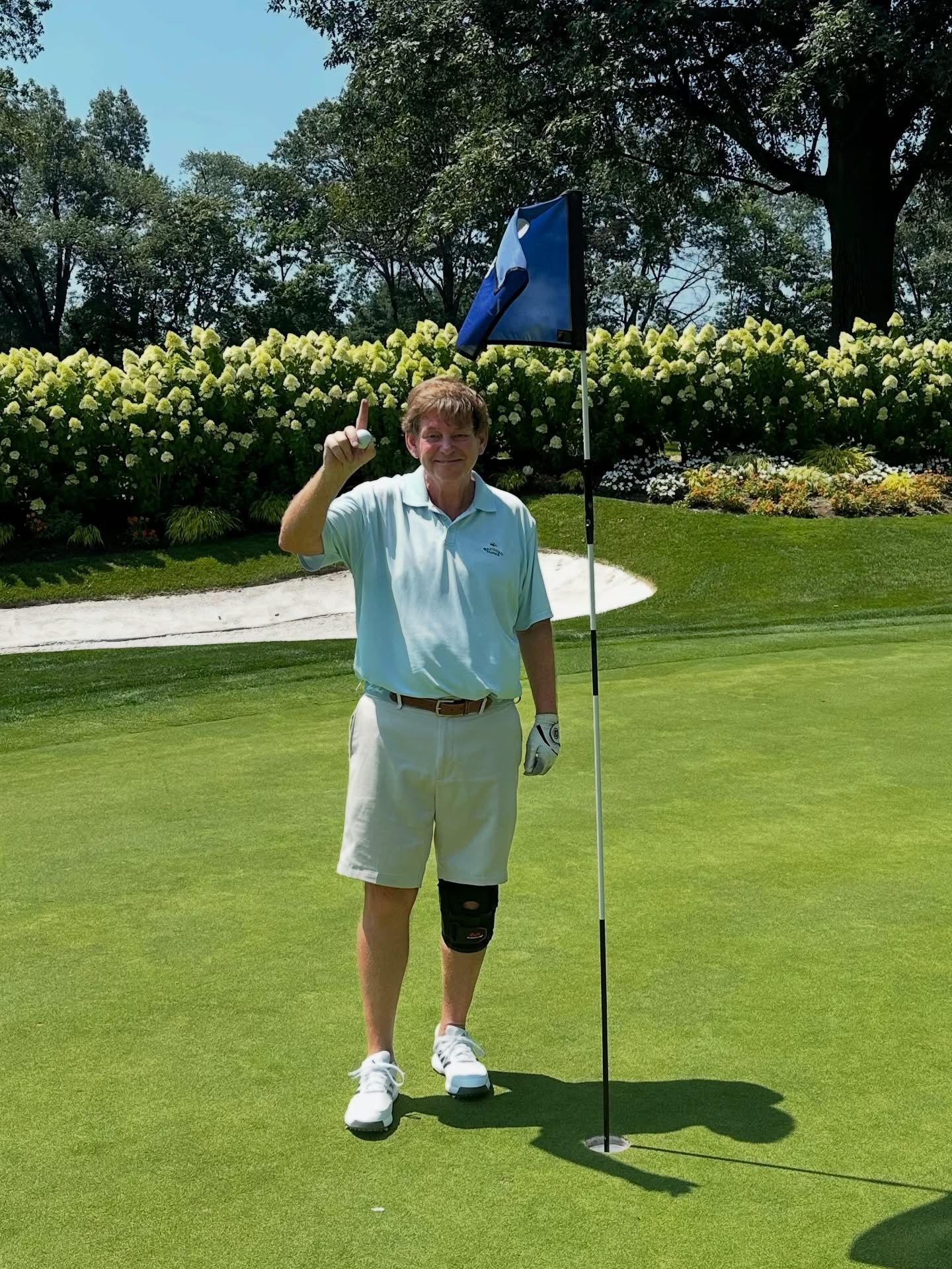 Man on golf green with blue flag, raising one finger. He's wearing a light blue shirt, khaki shorts, and a knee brace.