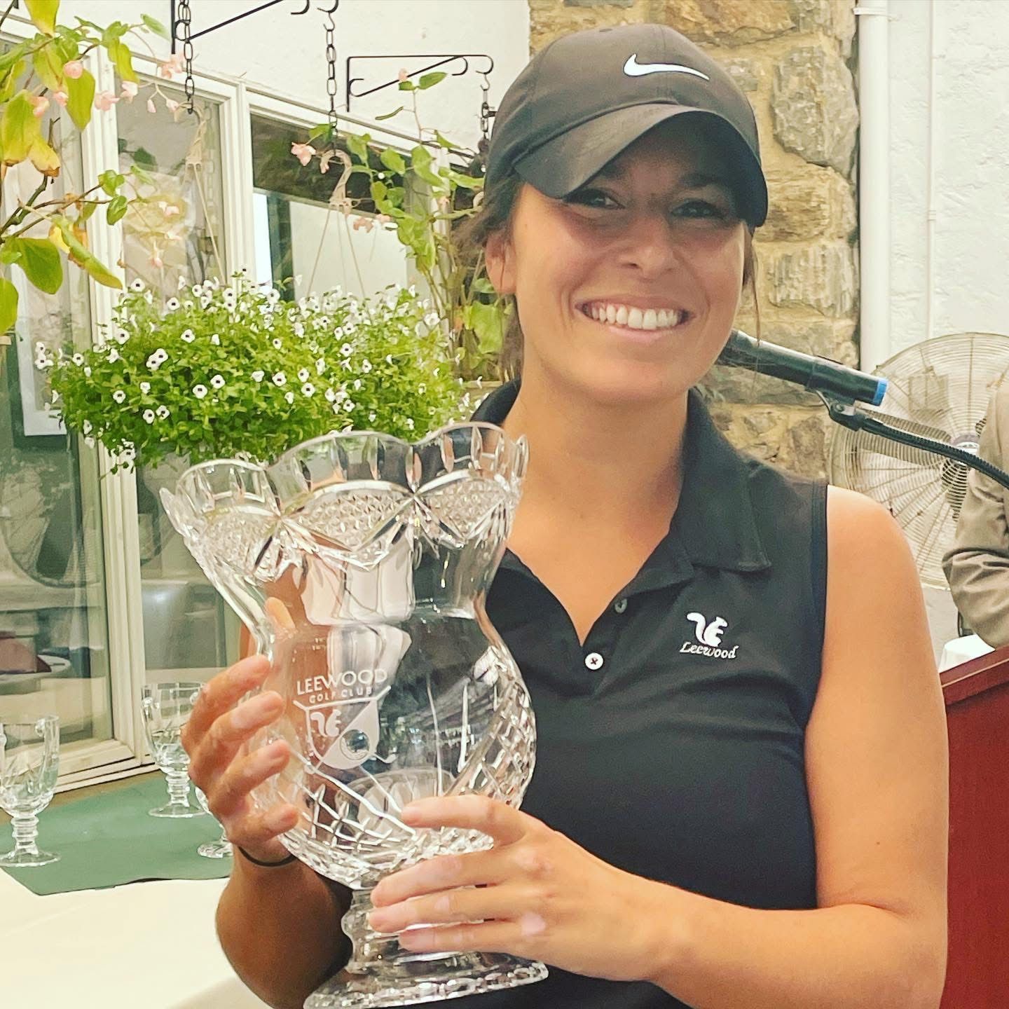 Woman smiles, holding a trophy outdoors, wearing a golf cap and polo shirt.