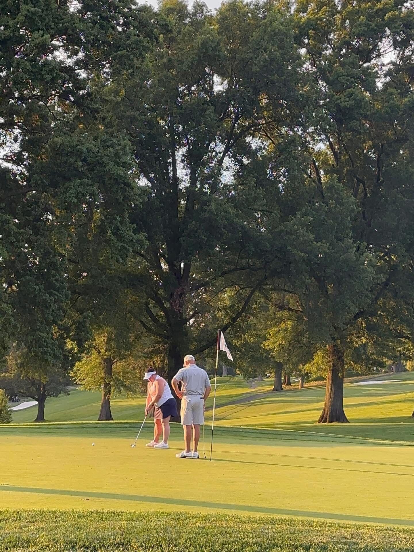 Two people putting on a golf course green, trees in the background, flag waving.