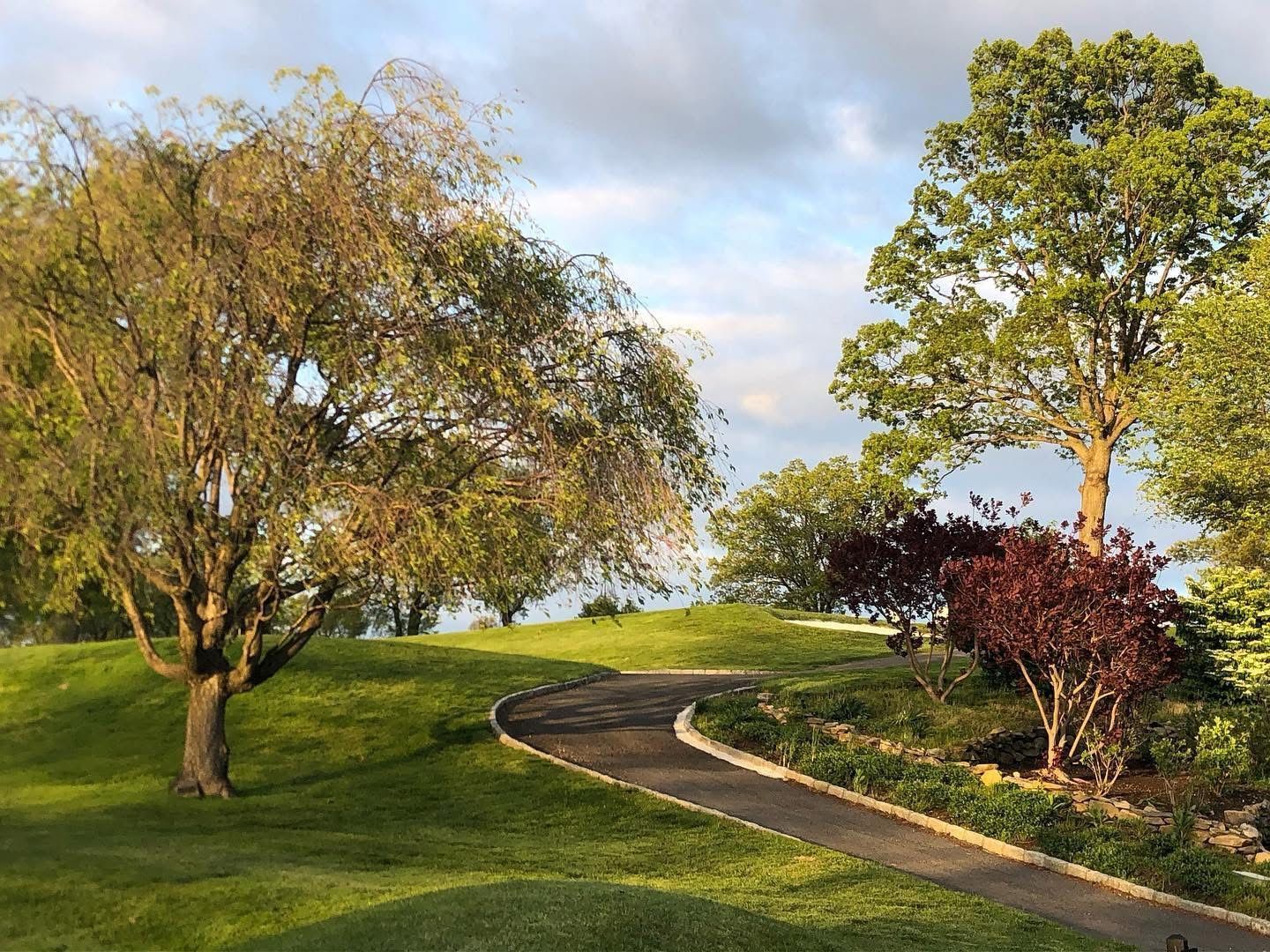 Path winds through a grassy hill, with trees and a cloudy sky in the background.