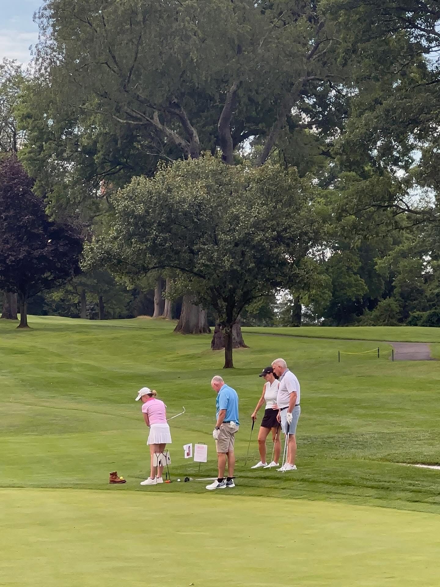 Golfers on a green course, preparing to tee off. One woman tees up, others watch, surrounded by trees.