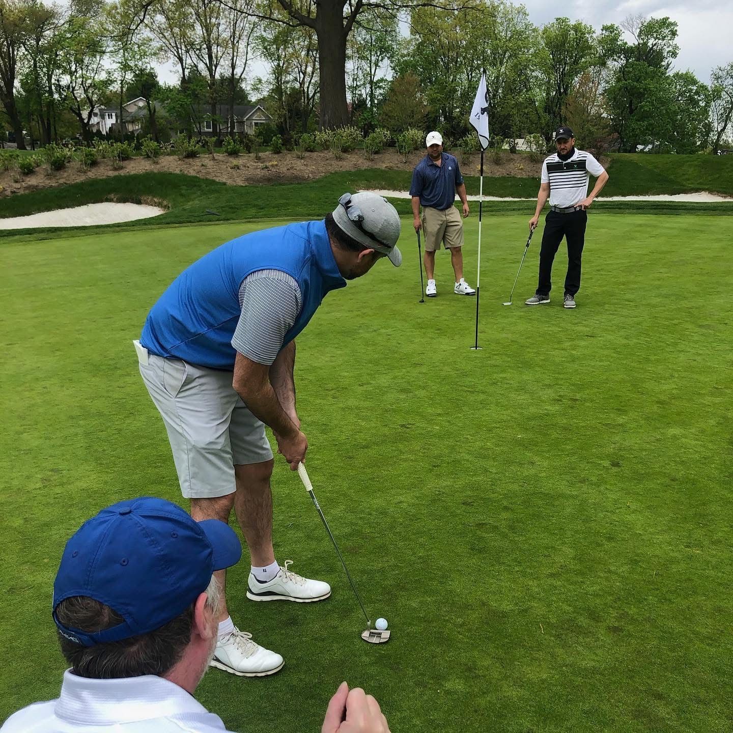 Men playing golf on a green. One putts, two watch, and flag in the background on a sunny day.