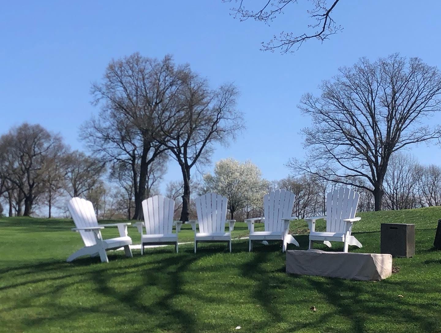 White Adirondack chairs on a grassy hill, trees in the background, bright blue sky.