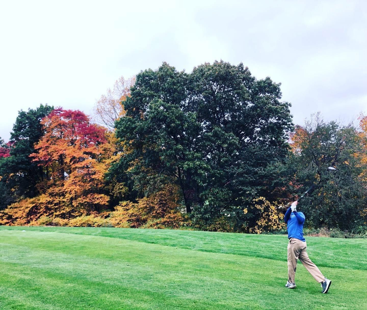 Golfer swinging club on green grass, colorful autumn trees in background. Cloudy sky.