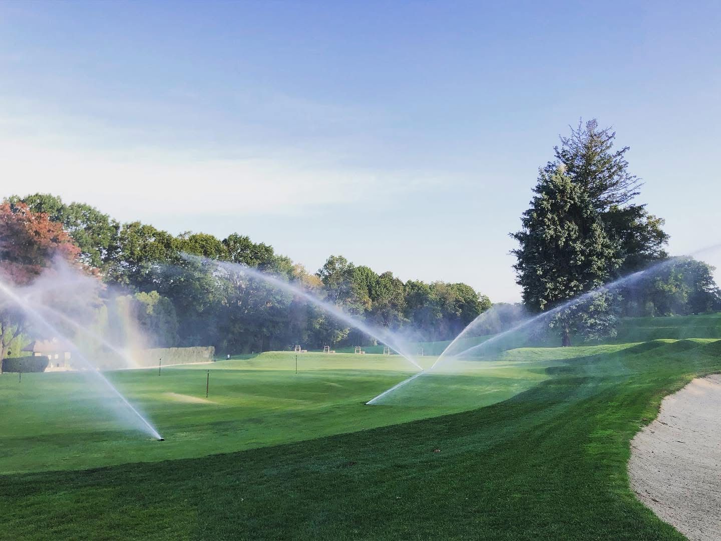 Sprinklers spraying water on a green golf course, with trees and blue sky in the background.