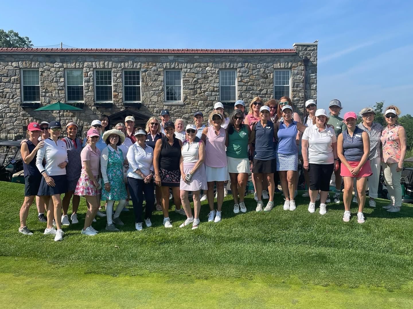 Group of golfers posing for a photo in front of a stone building on a sunny day.