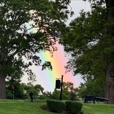 Rainbow over a golf course, framed by trees. A person stands on the green, with a black sign and a cart visible.