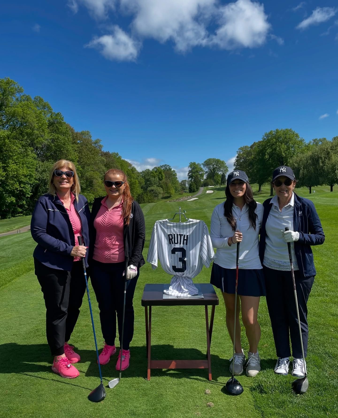 Four women on a golf course with a jersey. Blue skies, trees, and green grass. Smiling, holding golf clubs.