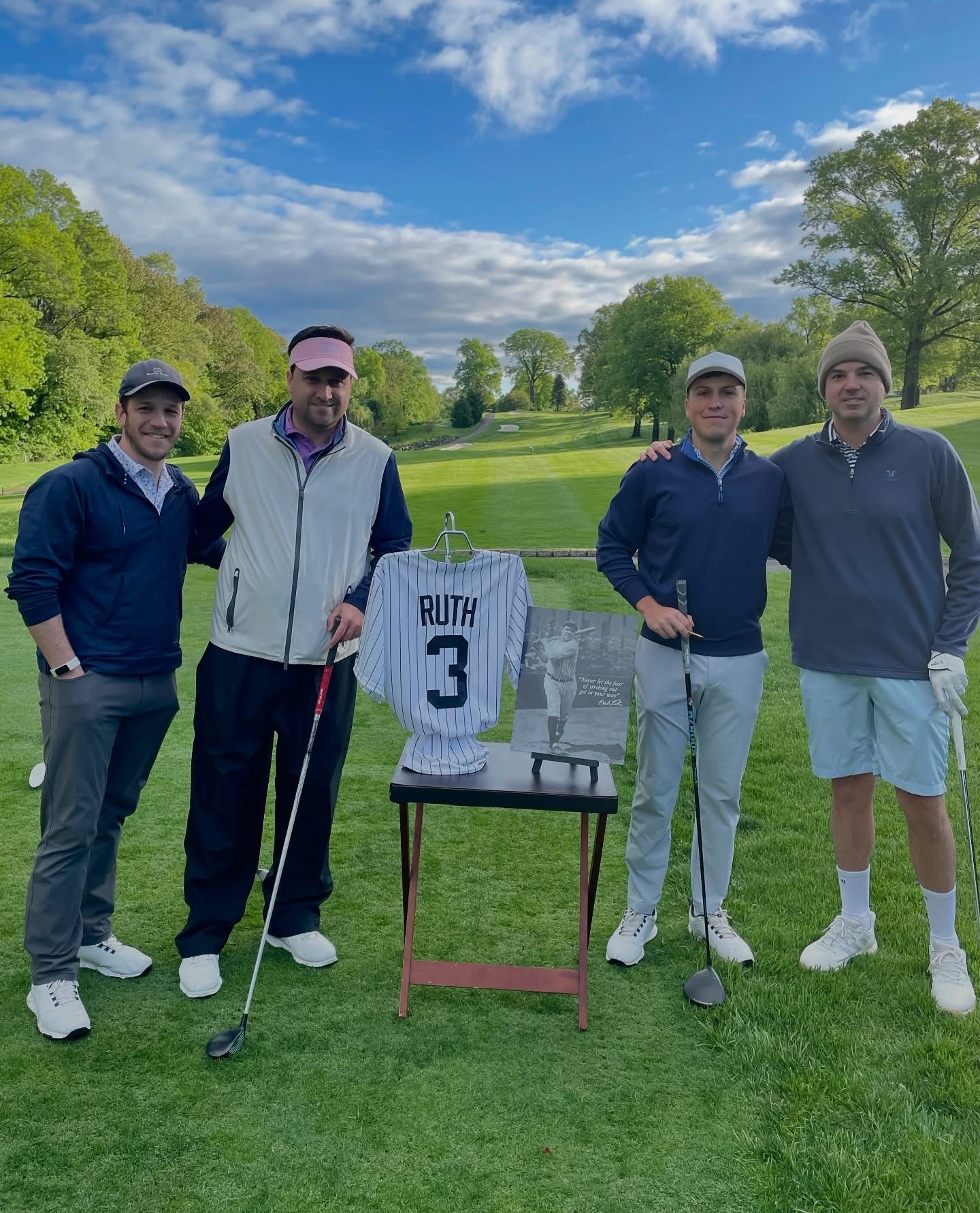 Four men on a golf course posing with a Babe Ruth jersey. Green grass, blue sky, and trees in the background.
