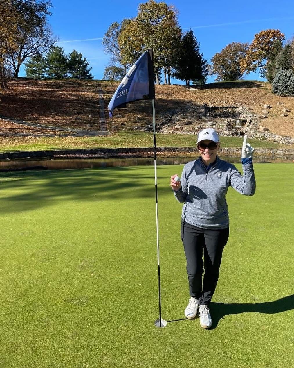 Woman on a golf course holds up one finger, smiling, near a flag and green grass.