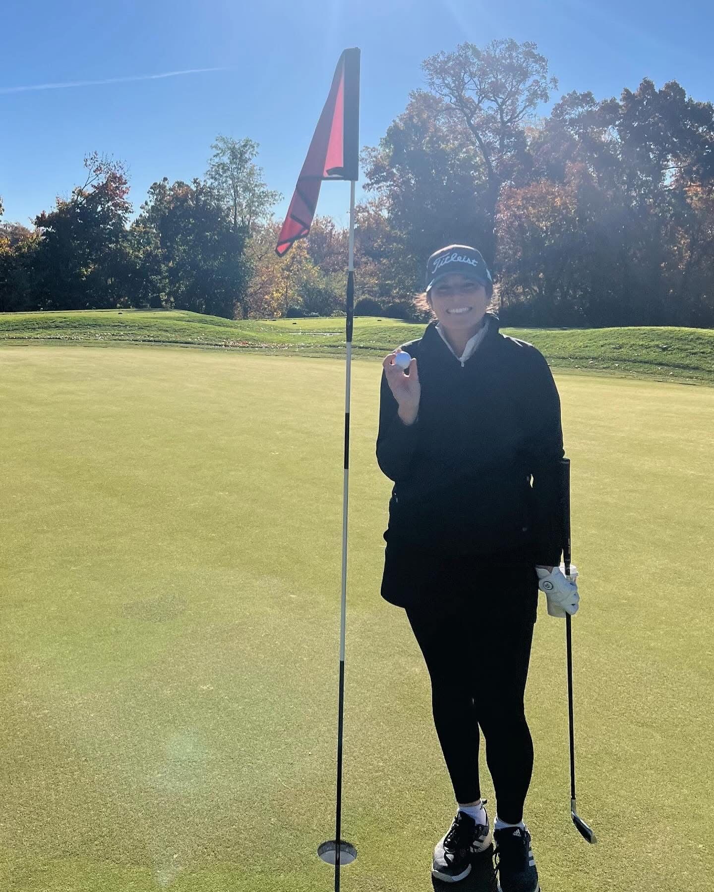 Woman on a golf course holds a ball near the hole, smiling. Red flag waves. Sunny day with green grass and trees.