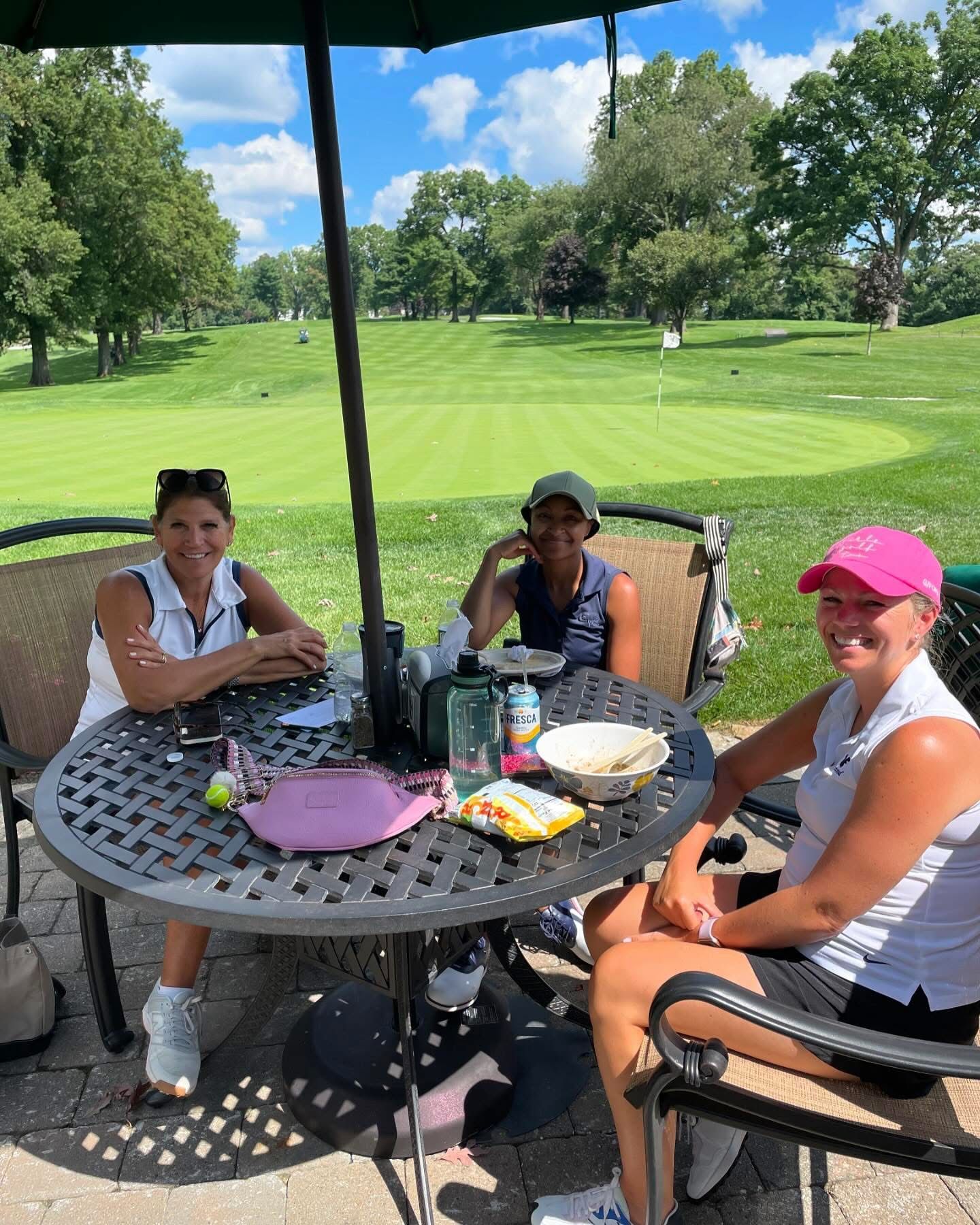 Three women sit at a patio table, golf course in background. Sunny day.