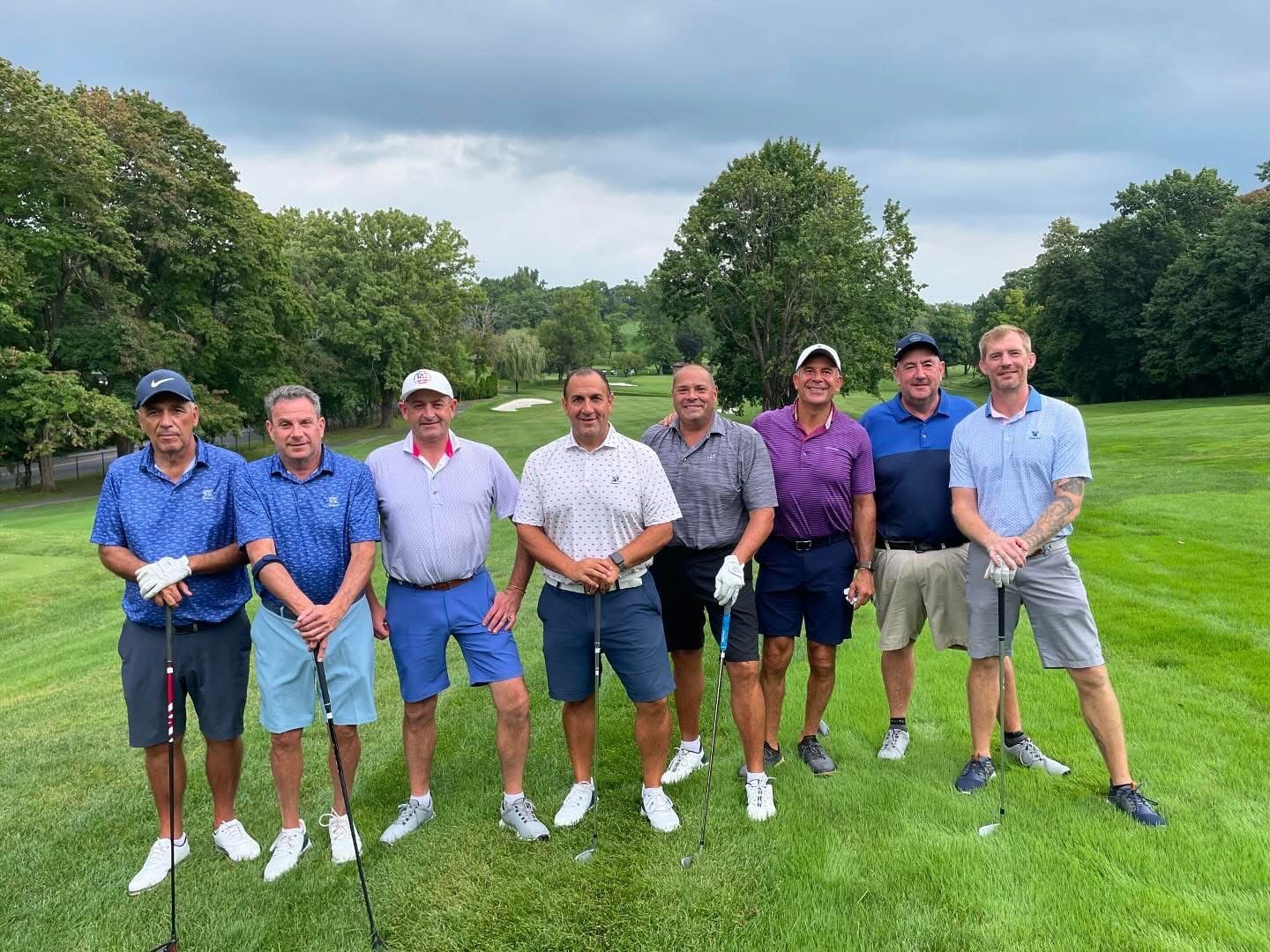 Group of men on a golf course, posing with clubs. They wear golf attire in various colors, trees and green grass visible.