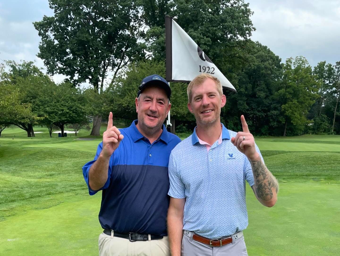 Two men on a golf course, pointing up. They wear golf attire. Green grass and a flag are in the background.