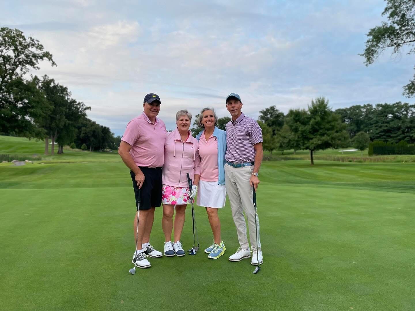 Four people in pink golf attire stand on a green, holding clubs.