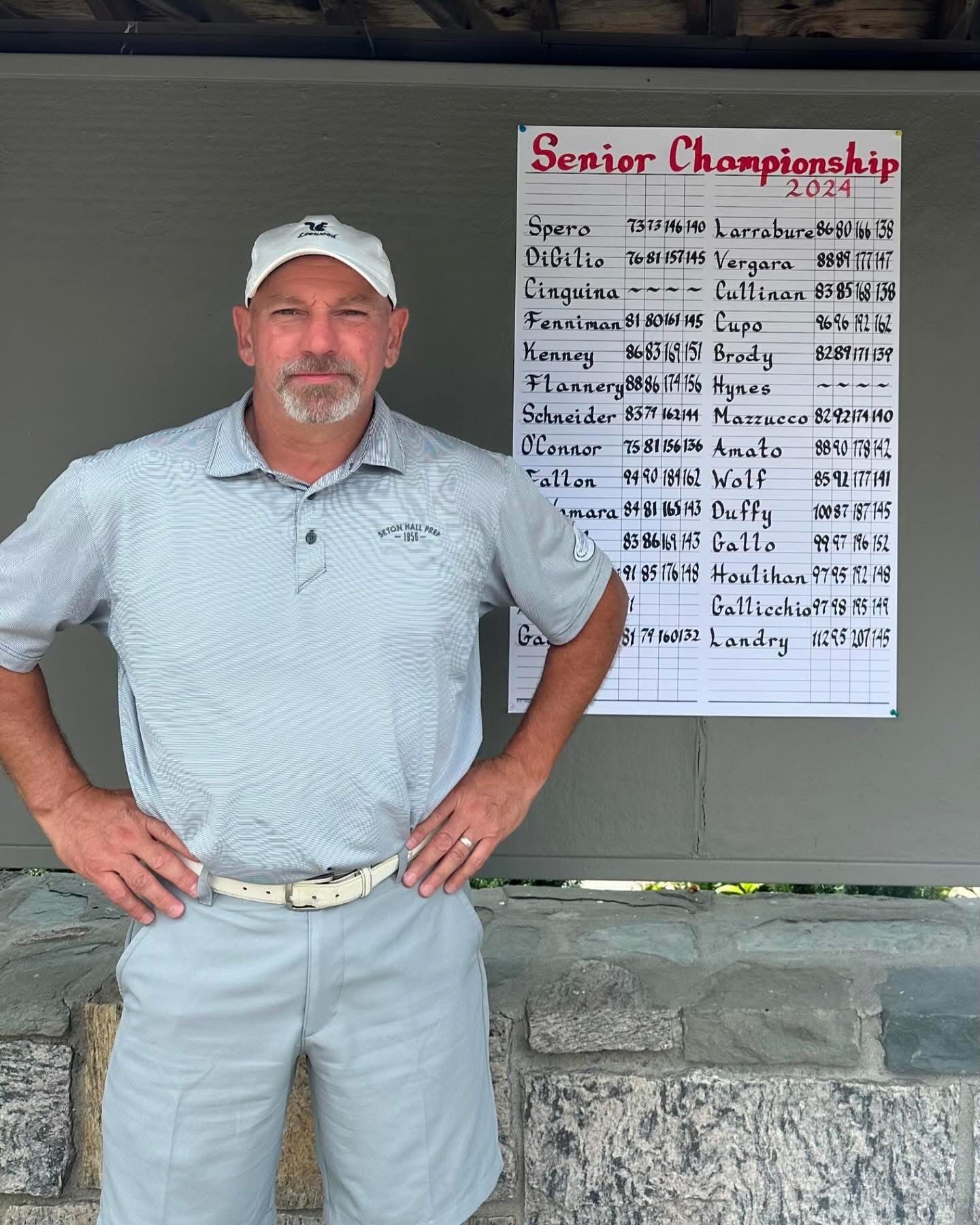 Man in golf attire stands near a scoreboard for a senior championship.