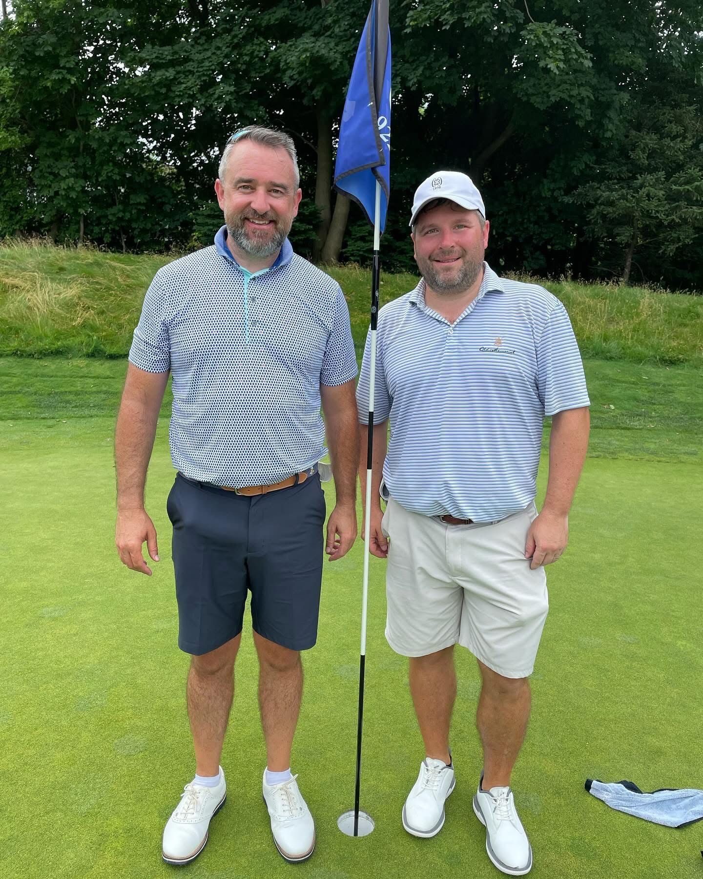 Two men in golf attire stand on a green next to a flag; one man's arm on the other's shoulder.