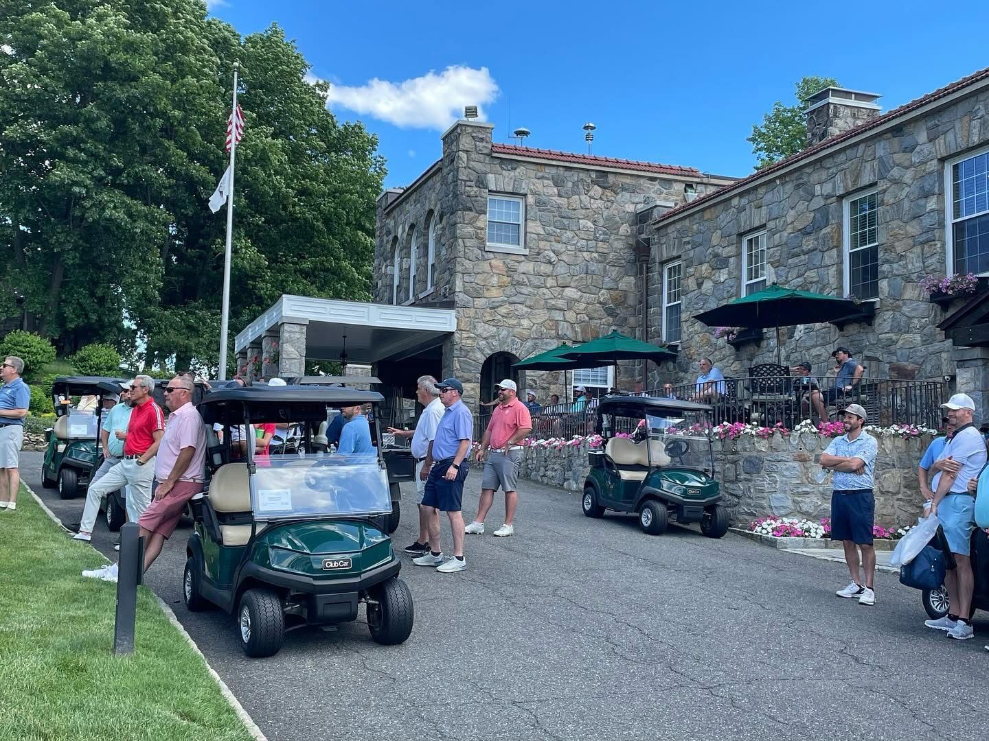 People in golf attire standing near golf carts, stone building with flag and umbrella.