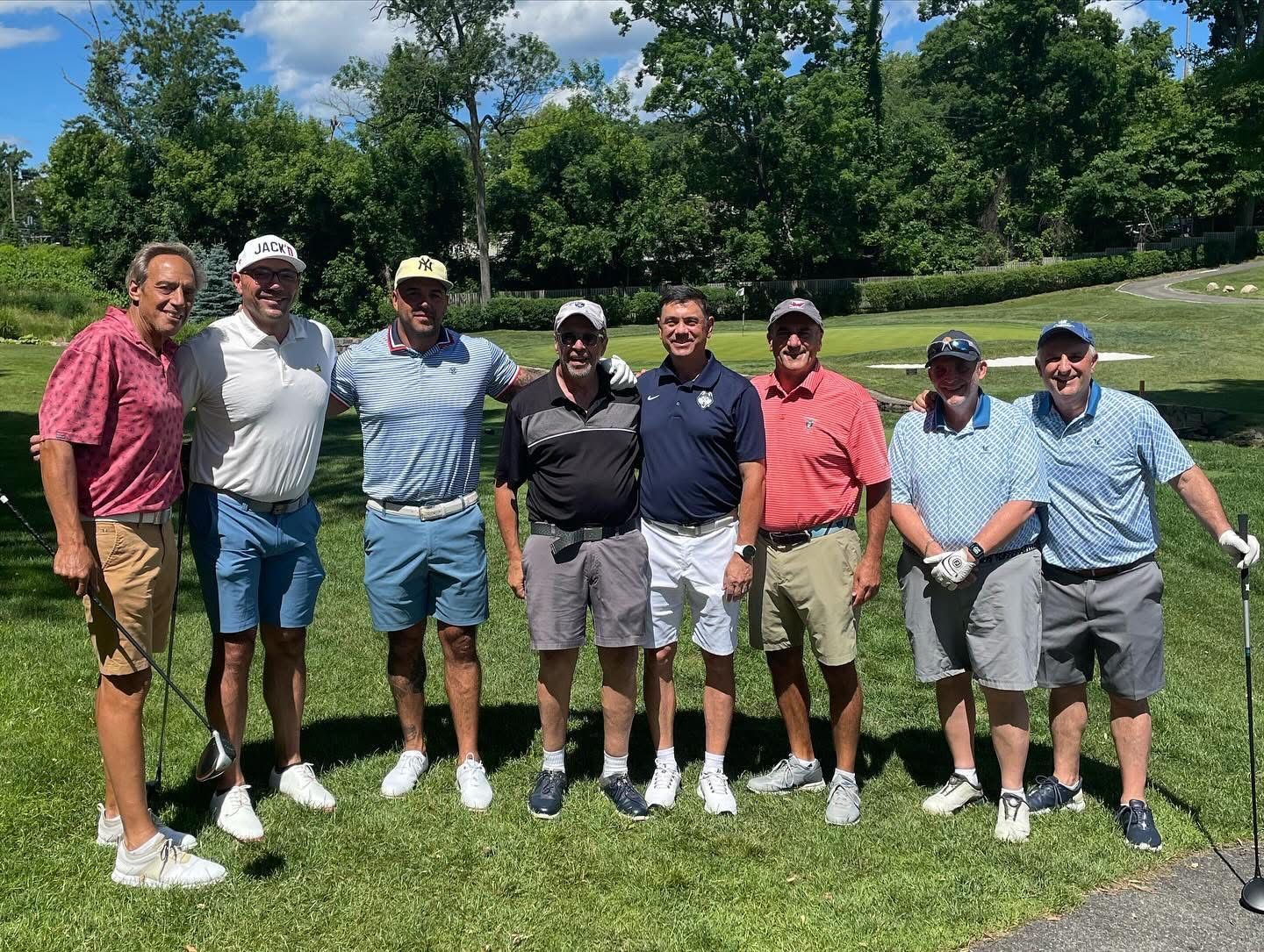 Group of golfers posing outdoors on a sunny golf course; men in golf attire smile, some with arms around each other.