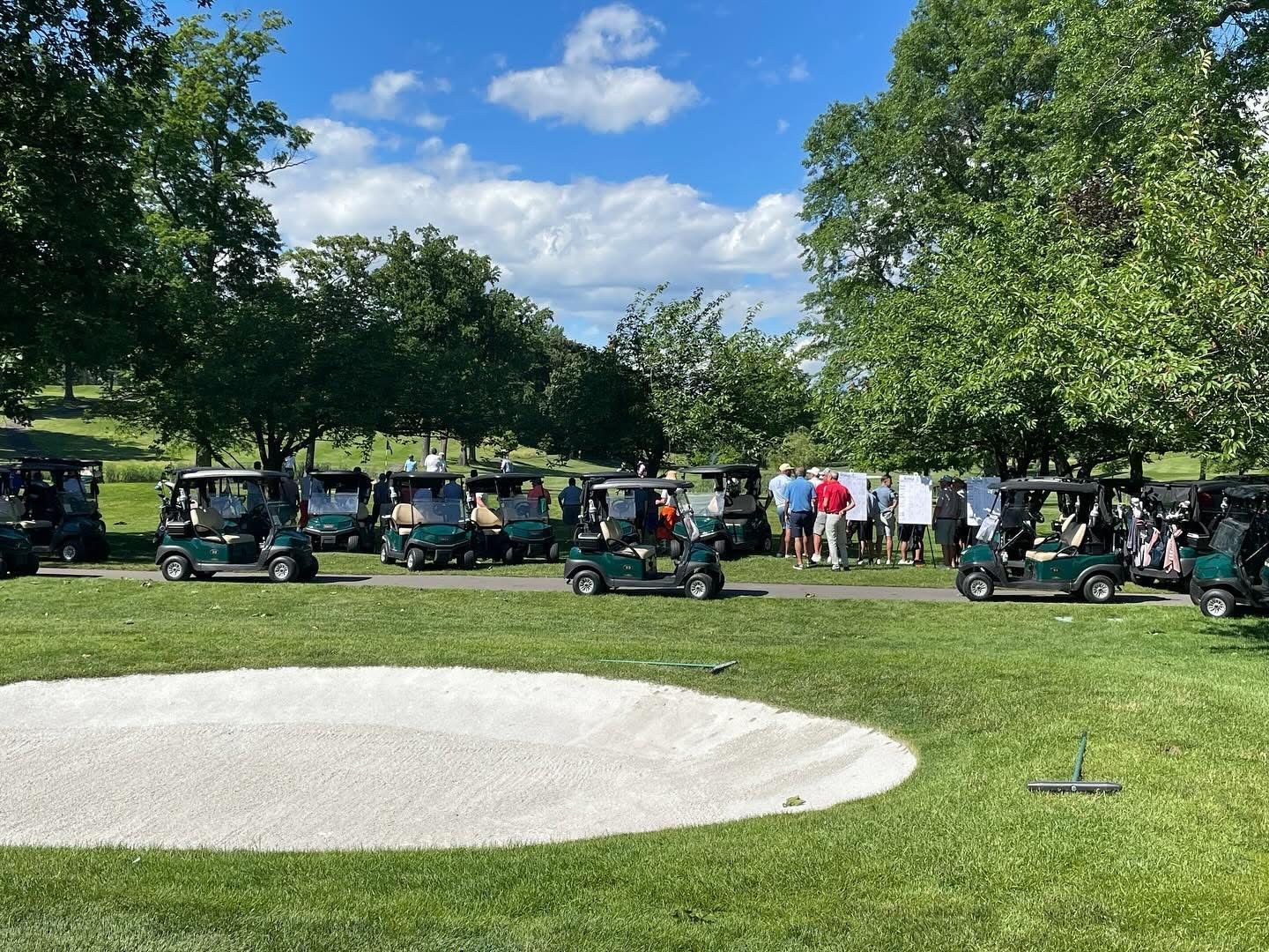 Golf carts parked on a green, near a sand trap, with people gathered in the background under a blue sky.