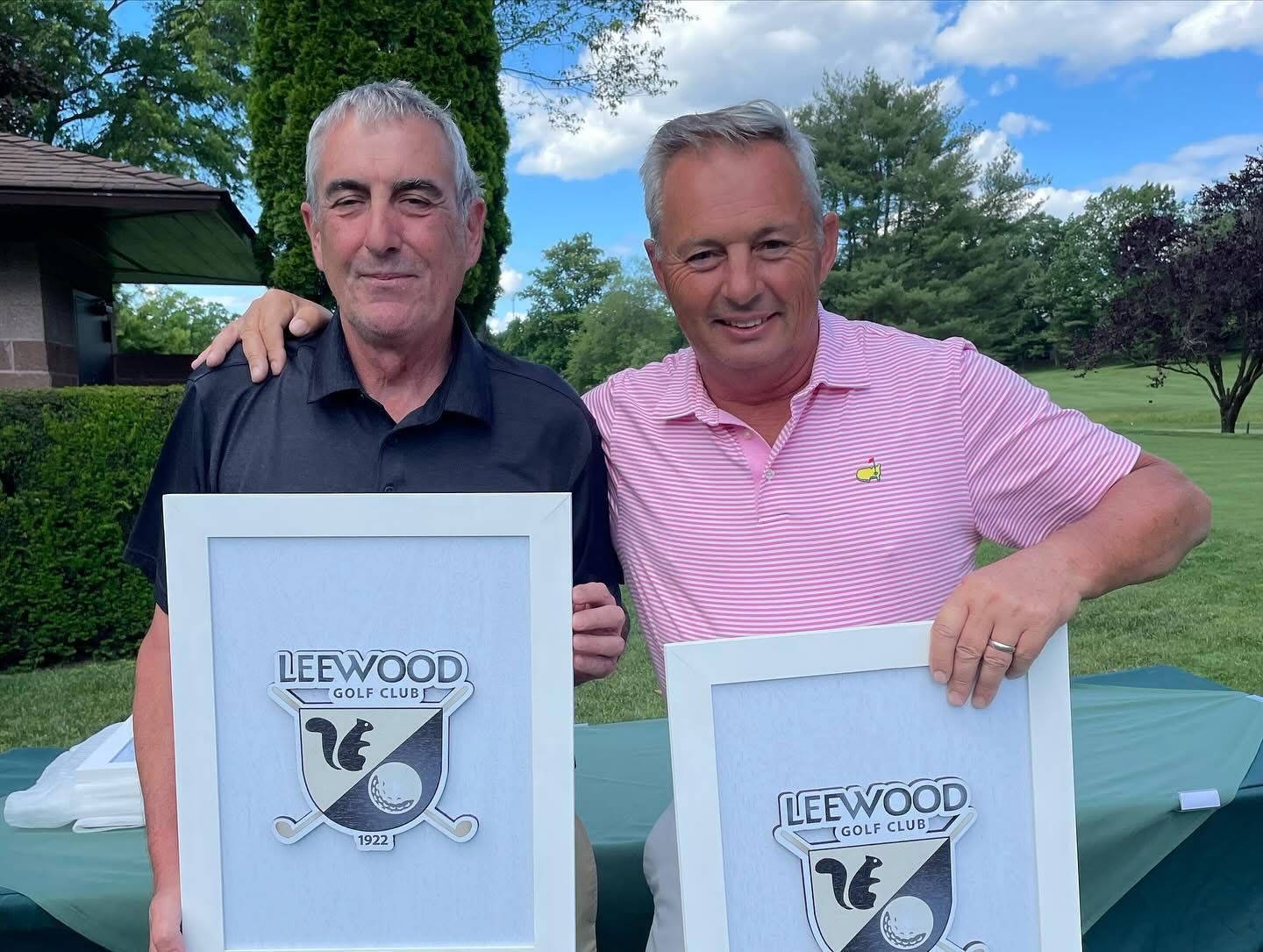 Two men holding framed logos, smiling outdoors on a golf course, with trees in the background.