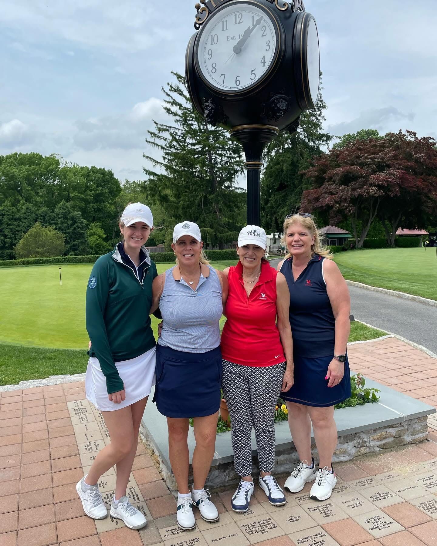 Four women golfers pose by a clock. They wear golf attire and stand on brick. The clock is tall and dark.