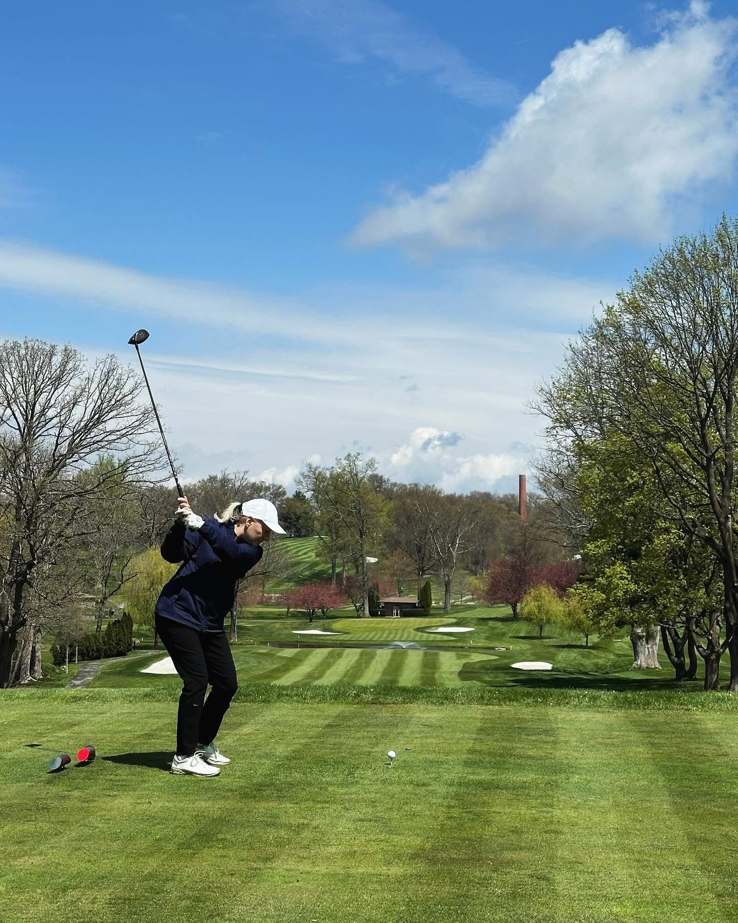Golfer swings club on a green golf course under a blue sky.