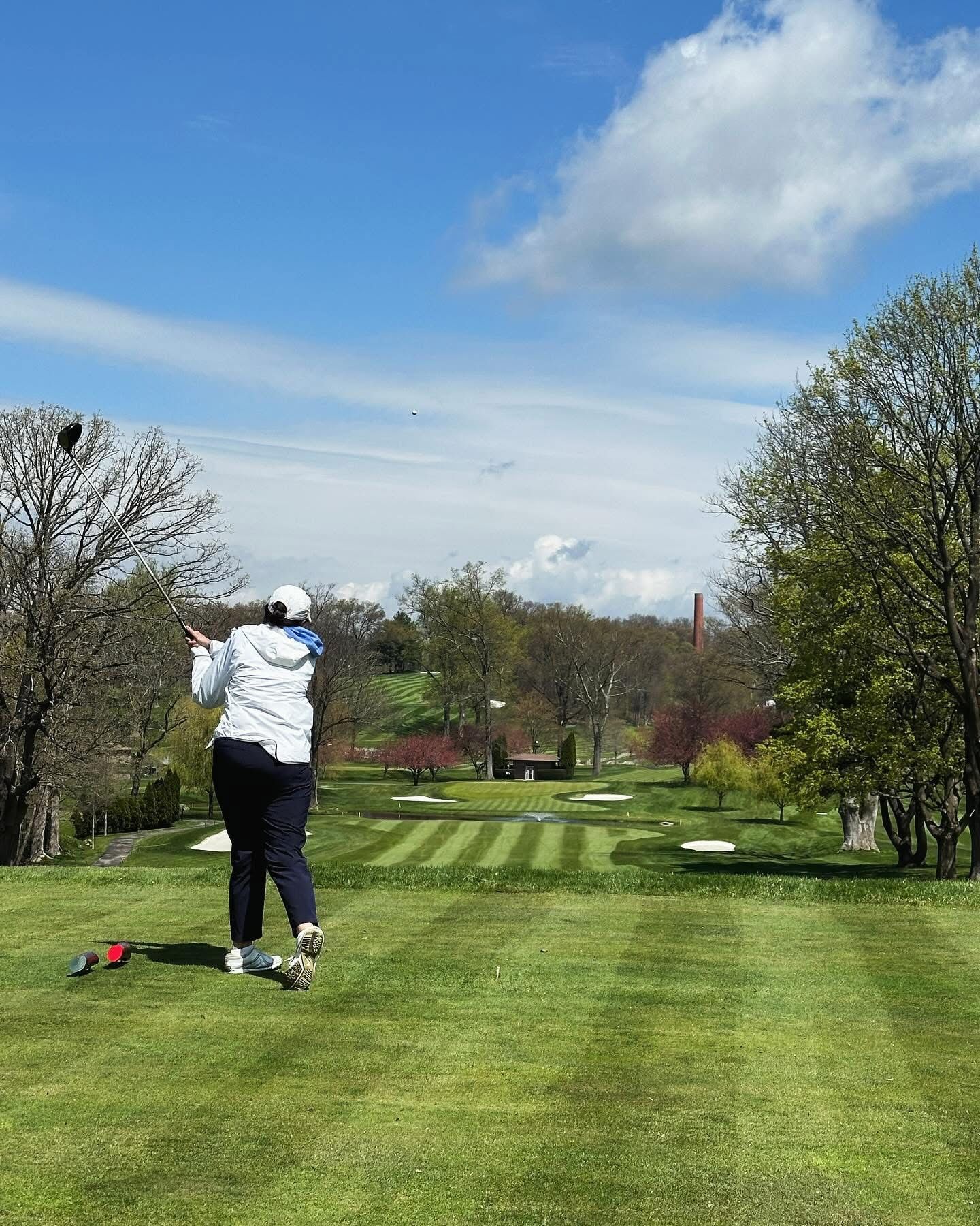 Golfer swings a club on a golf course under a blue sky, tee box in foreground, fairways in the distance.