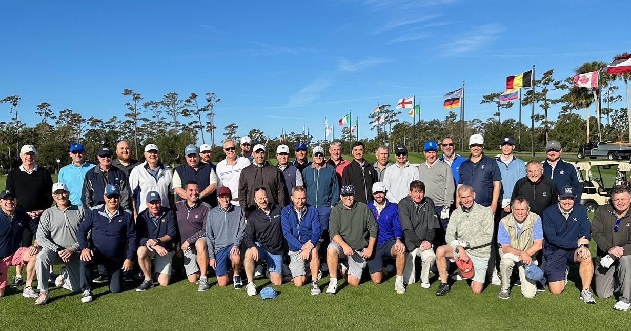 Group of men on a golf course posing for a photo under a sunny, clear sky; flags in the background.