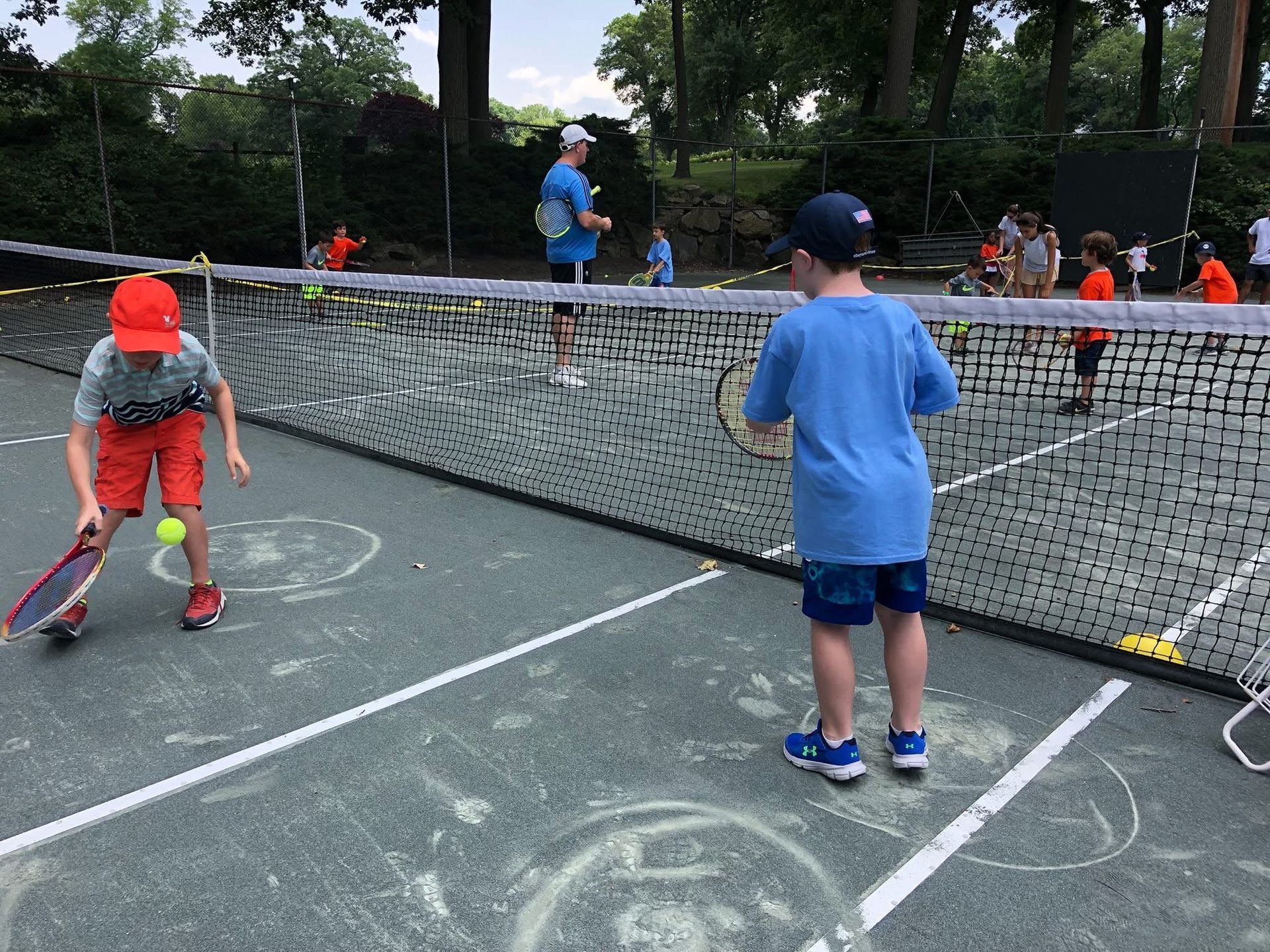 Children practice tennis on an outdoor court with a low net, hitting balls near painted targets on the ground.