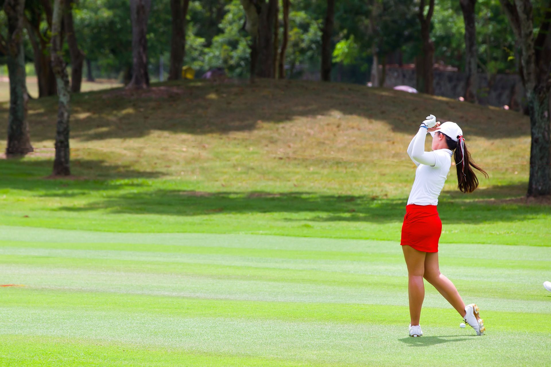 Woman in red skirt and white top swings a golf club on a green course.