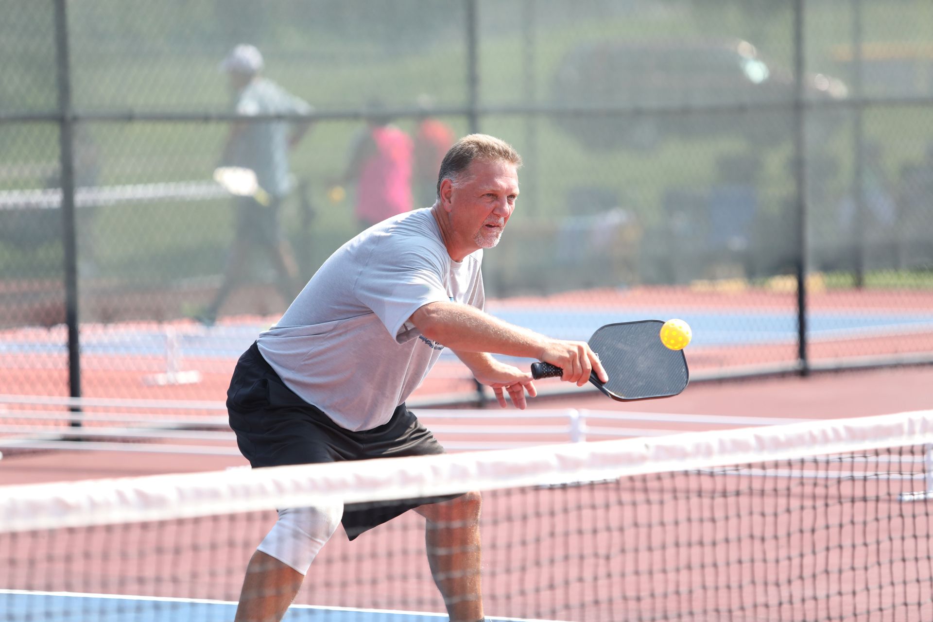 Man playing pickleball, hitting a yellow ball, near a net on a blue and red court.