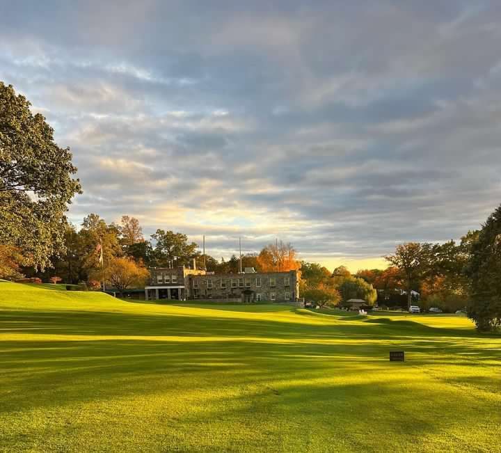 Large grassy lawn with stone building under a cloudy sky, trees in autumn colors.