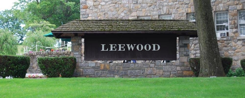 A wooden sign reading LEEWOOD mounted on a stone base in front of a stone building, surrounded by green grass and bushes.